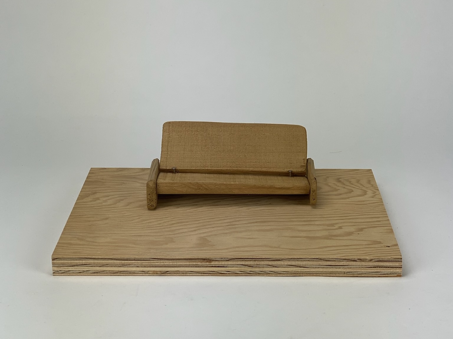 A model of a wooden couch sits atop a piece of plywood, against a white background.